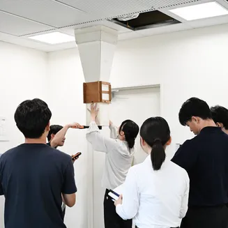 天井の吹出口に測定用フードを当てて風量を測定する学生たちの様子