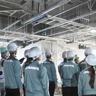 ヘルメットと作業服を着用した学生たちが、建設中の建物の天井に設置された配管を見上げている現場見学の様子