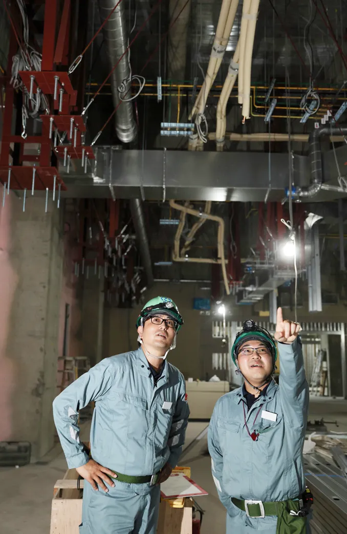 作業服とヘルメットを着用した社員2人が、施工中の天井に設置されたダクトや配管を見上げて指差している様子